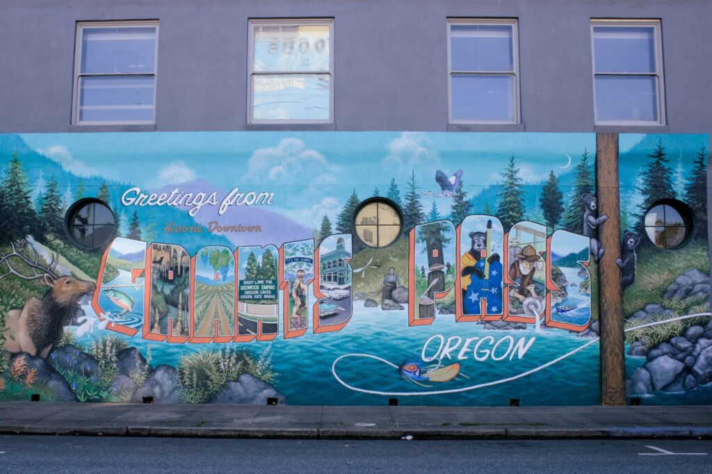 a mural that says "Greetings from historic downtown Grants Pass Oregon" The scene depicted in the mural is a nature scene with an elk, fly-fishing, and two black bears climbing trees. The letters in GRANTS PASS each have a different scene of either human activities that interconnect with nature as well as a black bear playing a stand-up bass. There is a building with windows showing above the mural