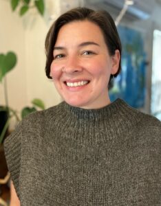 Head shot of Kathryn Duvall, who has brown hair, light skin, and a big smile and is wearing a brown sweater.